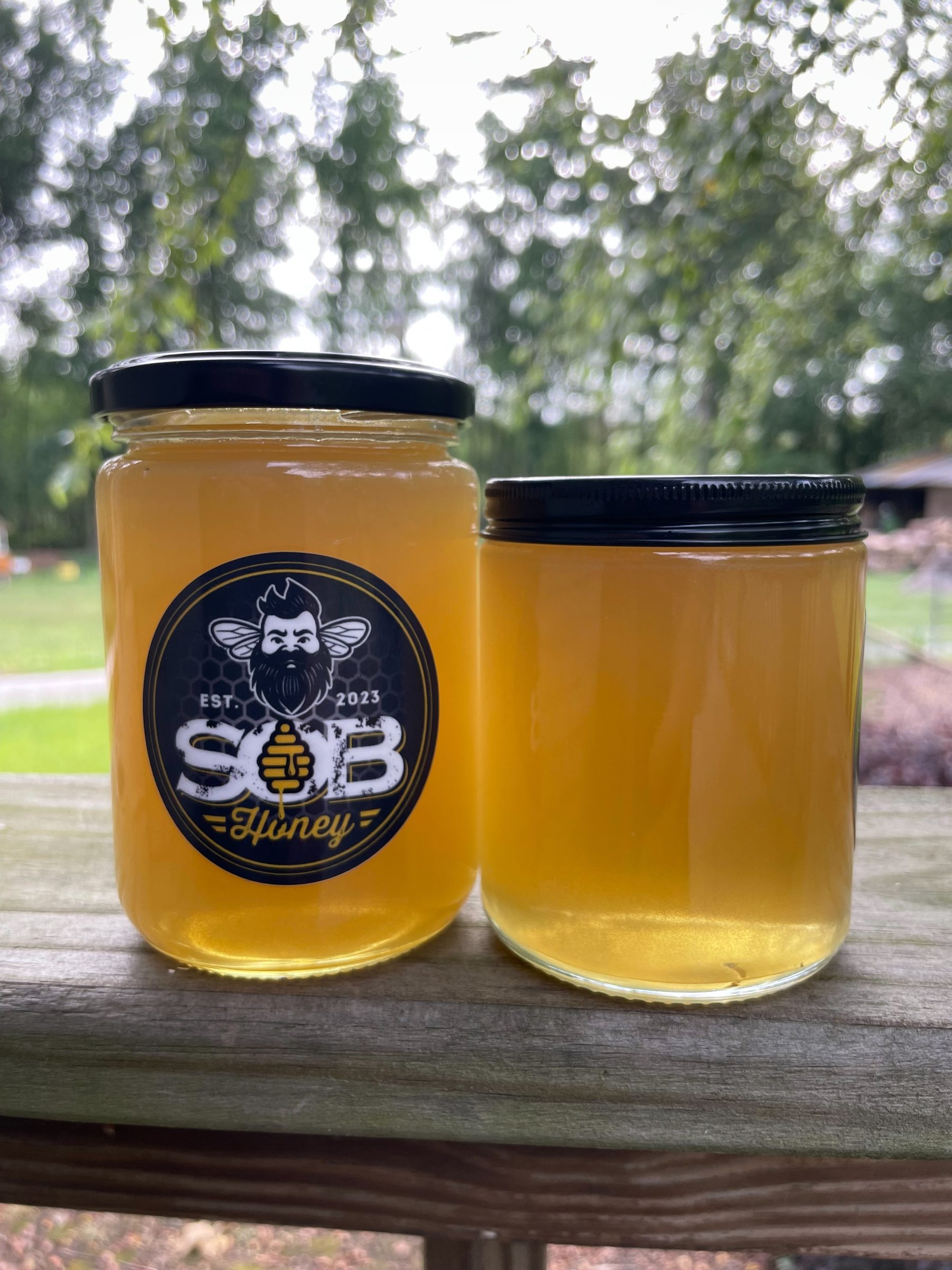Two jars of SOB Honey on a wooden rail at the yard, with the green of the trees behind them.
